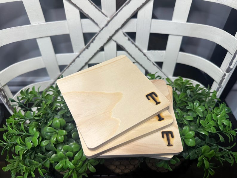 Puede incluir: Tres posavasos de madera clara con la letra "T" grabada. Los posavasos est&aacute;n apilados sobre plantas verdes, con una cesta blanca de fondo. Cada posavasos mide aproximadamente 10 cm.