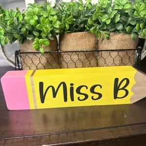 May include: A wooden pencil-shaped desk sign painted yellow with a pink eraser end and a tan tip. The text "Miss B" is printed in black script lettering across the yellow body. The pencil features a metallic-colored band between the eraser and body.