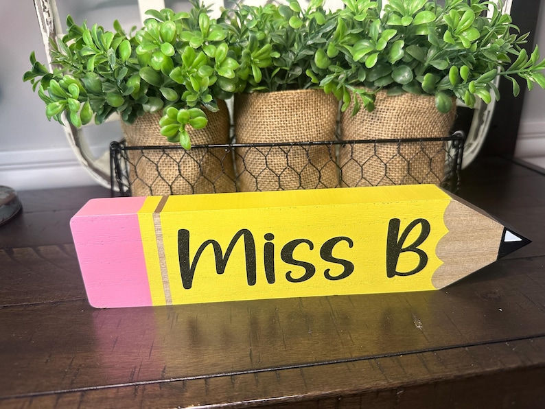 May include: A wooden pencil-shaped desk sign painted yellow with a pink eraser end and a tan tip. The text "Miss B" is printed in black script lettering across the yellow body. The pencil features a metallic-colored band between the eraser and body.