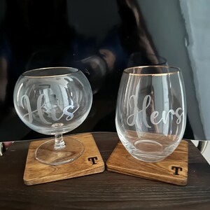 Puede incluir: Dos copas de vino transparentes con bordes dorados, una con la inscripci&oacute;n "His" y la otra "Hers". Se apoyan en posavasos cuadrados de madera con la letra "T". Las copas est&aacute;n sobre una superficie de madera oscura.