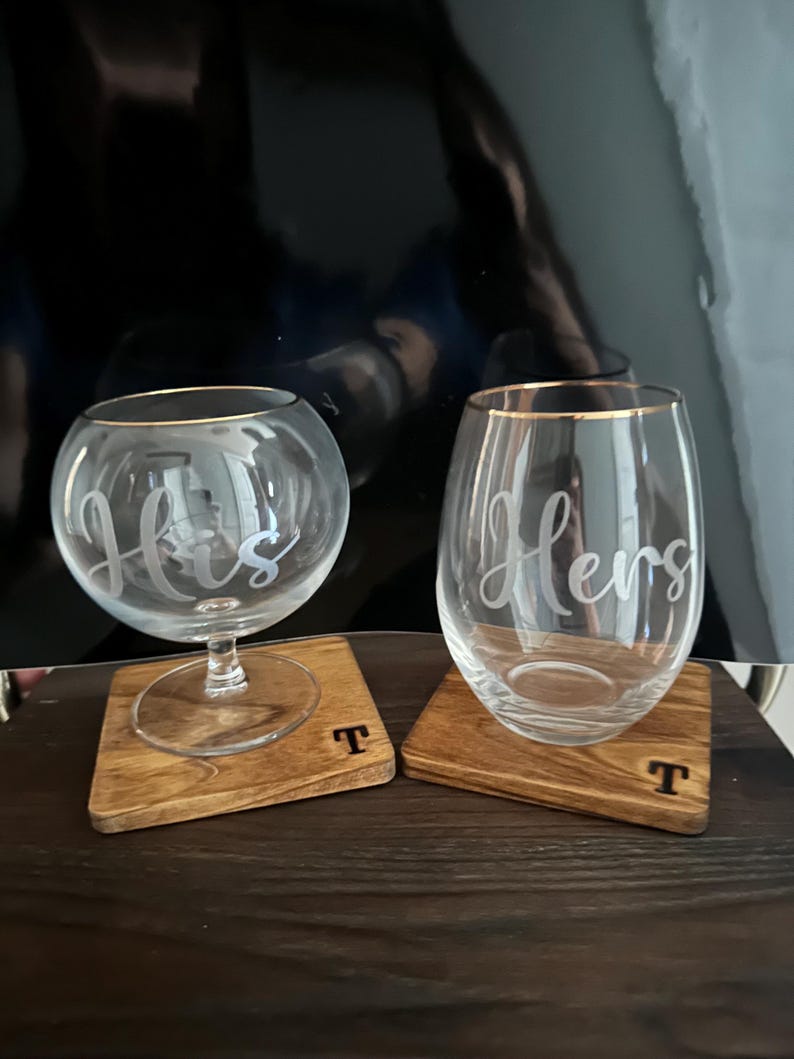 Puede incluir: Dos copas de vino transparentes con bordes dorados, una con la inscripci&oacute;n "His" y la otra "Hers". Se apoyan en posavasos cuadrados de madera con la letra "T". Las copas est&aacute;n sobre una superficie de madera oscura.