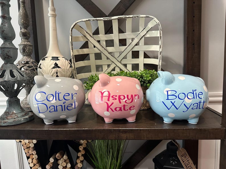 May include: Three ceramic piggy banks in gray, pink, and blue, each with white polka dots. The gray bank has "Colter Daniel" in blue script, the pink bank has "Aspyn Kate" in pink script, and the blue bank has "Bodie Wyatt" in blue script.