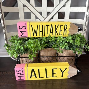 May include: Two wooden pencil-shaped signs with yellow bodies, pink erasers, and brown tips. The top sign reads MS. WHITAKER in black lettering, and the bottom sign reads MRS. ALLEY in black lettering. Both signs are positioned in front of green foliage.