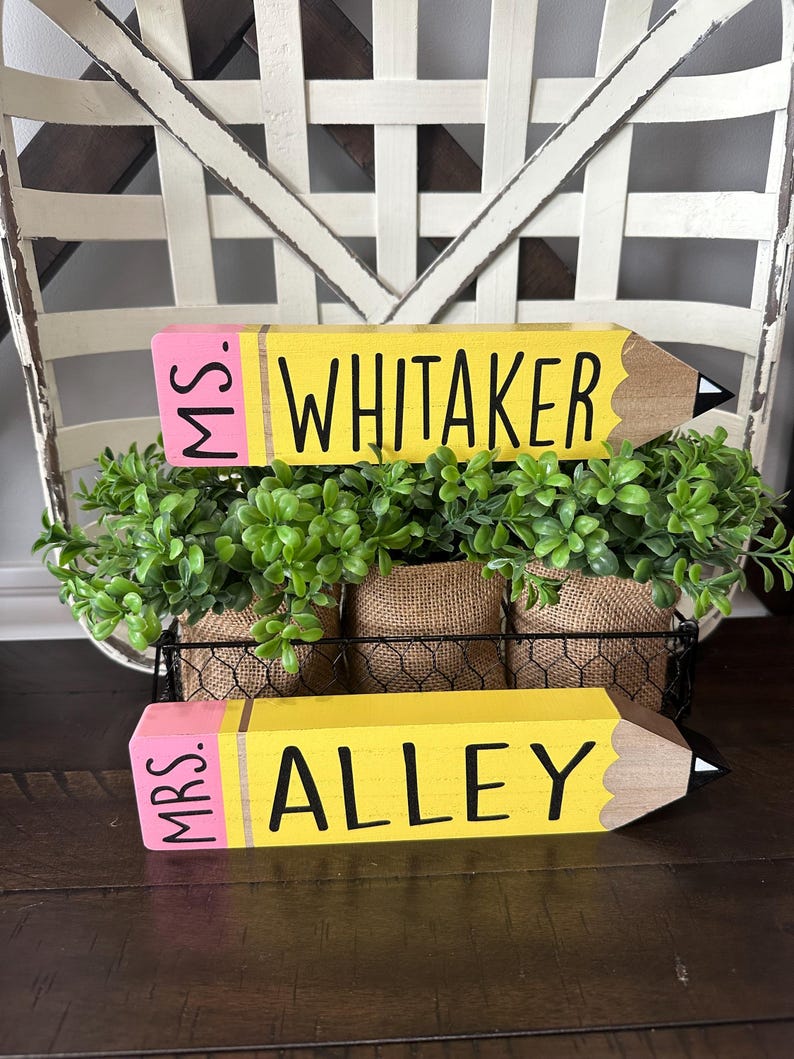 May include: Two wooden pencil-shaped signs with yellow bodies, pink erasers, and brown tips. The top sign reads MS. WHITAKER in black lettering, and the bottom sign reads MRS. ALLEY in black lettering. Both signs are positioned in front of green foliage.