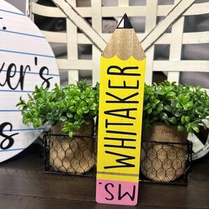 May include: A wooden pencil-shaped sign featuring a yellow body with the name WHITAKER printed in black, a tan wood-grain tip with a black point, and a pink eraser base with the text MS. printed in black on the pink section.