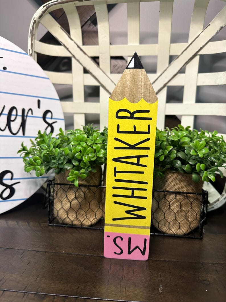 May include: A wooden pencil-shaped sign featuring a yellow body with the name WHITAKER printed in black, a tan wood-grain tip with a black point, and a pink eraser base with the text MS. printed in black on the pink section.