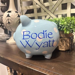 May include: A light blue ceramic piggy bank with white polka dots. The name "Bodie Wyatt" is written in blue script across the front. The piggy bank has four small feet and a small tail.