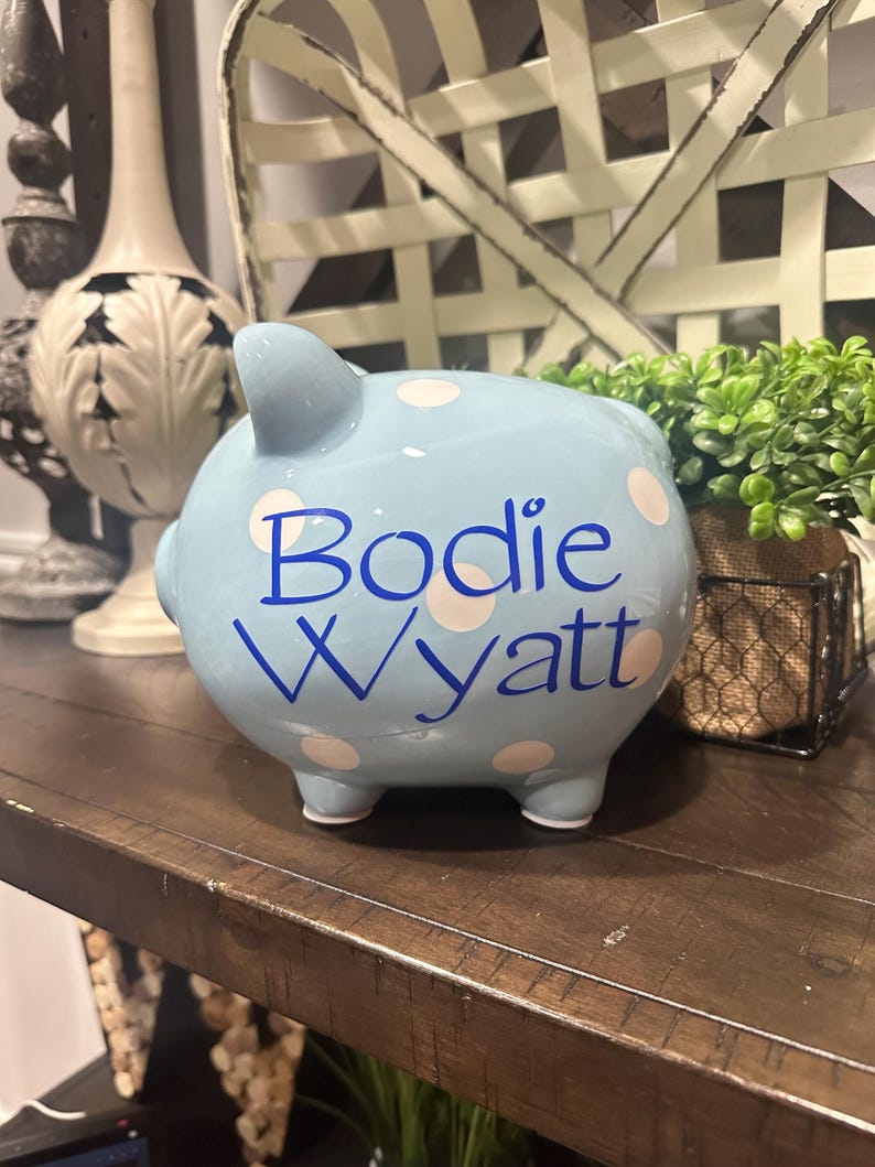 May include: A light blue ceramic piggy bank with white polka dots. The name "Bodie Wyatt" is written in blue script across the front. The piggy bank has four small feet and a small tail.