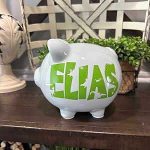 Peut inclure: Une tirelire en c&eacute;ramique blanche avec le nom "ELIAS" en lettres vert vif et des silhouettes de dinosaures. La tirelire a quatre petits pieds et une fente sur le dessus pour ins&eacute;rer des pi&egrave;ces. Elle est pos&eacute;e sur une surface en bois fonc&eacute;.