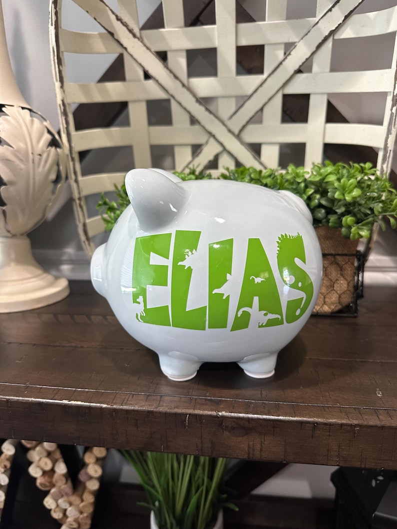 Peut inclure: Une tirelire en c&eacute;ramique blanche avec le nom "ELIAS" en lettres vert vif et des silhouettes de dinosaures. La tirelire a quatre petits pieds et une fente sur le dessus pour ins&eacute;rer des pi&egrave;ces. Elle est pos&eacute;e sur une surface en bois fonc&eacute;.