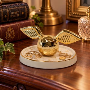 May include: A gold-colored spherical trinket dish with outstretched wings, centered on a round white ceramic tray featuring gold star patterns. The tray holds two gold rings, one with a clear stone, and a delicate gold chain with a circular pendant.