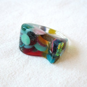 Pode incluir: Um anel de resina colorido com uma variedade de materiais embutidos, incluindo glitter, pequenas contas e flocos iridescentes. O anel é uma peça de joia única e chamativa.