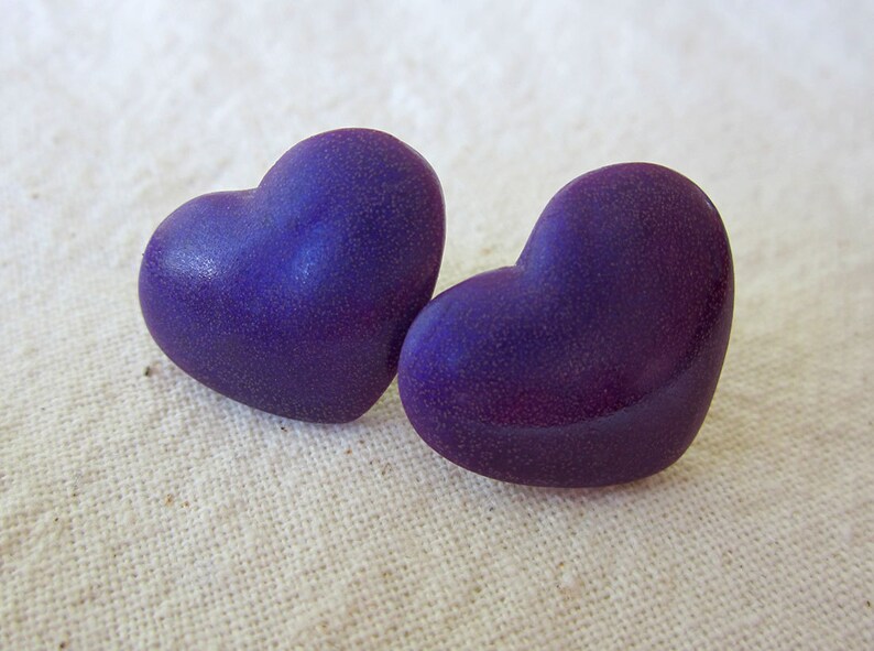 Puede incluir: Dos pendientes de color morado en forma de coraz&oacute;n con una superficie texturizada.