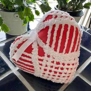 May include: A red and white heart-shaped pillow with a white bow. The pillow is made of a soft, fluffy material and has a textured surface.