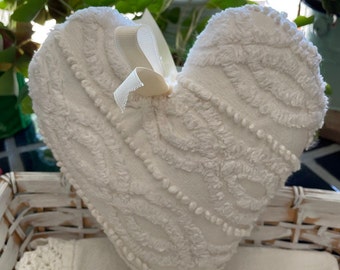 May include: White heart-shaped pillow with a textured surface and a white satin ribbon bow. The pillow is sitting in a white wicker basket.