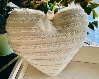 May include: A white heart-shaped pillow with a white ribbon. The pillow has a textured, fluffy surface with horizontal stripes.