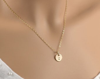 May include: A delicate gold-colored necklace with a small, round pendant. The pendant features the letter "S" in the center. The necklace is displayed on a person wearing a white top with pearl-like embellishments.
