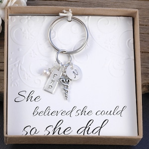 May include: A silver keychain with a charm of a medical caduceus, a small pearl, and a tag that says "FNP". The keychain is in a small box with a white background and the text "She believed she could so she did".
