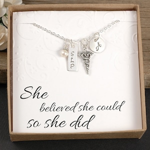 May include: A silver necklace with multiple charms, including a pearl, a rectangular tag with "SLP", a medical symbol, and a round charm with the letter "A". The necklace is presented in a white box with the text "She believed she could so she did".