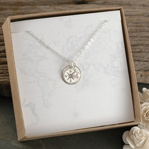 May include: A silver necklace with a compass pendant and a small pearl charm, presented in a gift box. The compass pendant features directional markings. The background includes a world map design.