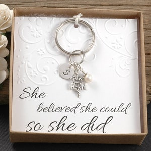 May include: A silver keychain with a charm that says "RN" and a small pearl. The keychain is in a box with a white card that says "She believed she could so she did".