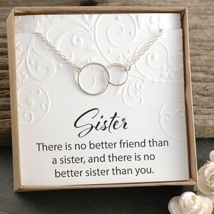 May include: A silver necklace with two interlocking circles, presented in a white box with a brown lid. The box has a white card with the text "Sister. There is no better friend than a sister, and there is no better sister than you."
