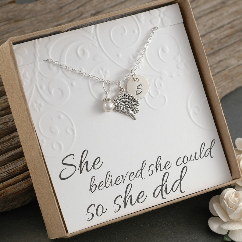 May include: A silver necklace with a brain charm, a pearl, and a silver disc with the letter "S" on it. The necklace is in a white box with the text "She believed she could so she did".