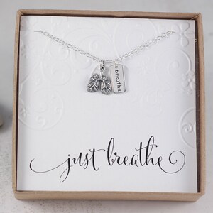 May include: A silver necklace with a charm of a pair of lungs and a tag that says "breathe". The necklace is in a white box with the words "just breathe" written in cursive.