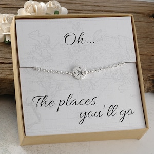 May include: A silver compass charm bracelet with a white card that reads "Oh... The places you'll go". The card is on a brown background.
