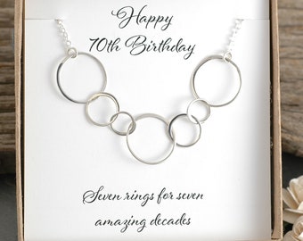 May include: A silver necklace with seven interconnected circles, presented in a white gift box with a card that reads "Happy 70th Birthday" and "Seven rings for seven amazing decades".