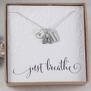 May include: A silver necklace with a charm that says "breathe" and a charm that looks like lungs. The necklace also has a small round charm with the letter "M" on it. The necklace is in a white box with the words "just breathe" written in cursive.