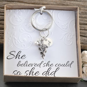 May include: A silver key chain with a snake and staff charm, a pearl bead, and a personalized disc with the letter "A". The keychain is in a small box with a white background and the text "She believed she could so she did".