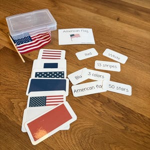 May include: Educational flashcards and a small American flag are displayed on a wooden surface. The flashcards feature the American flag and its components: red, white, and blue colors, 13 stripes, and 50 stars. A clear plastic container holds the flag.