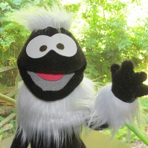May include: A black and white puppet with large white eyes and a red mouth. The puppet has white fur around its face and on its arms. The puppet is set against a blurred green background.