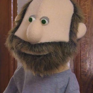 May include: A handmade puppet with a light beige head, a brown furry beard, and green eyes. The puppet is wearing a gray long-sleeved shirt. The puppet's head is bald on top and has brown fur around the sides.