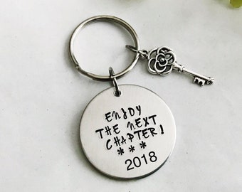 Puede incluir: Llavero de plata con un dije redondo que dice "Enjoy the next chapter! *** 2018". El dije está unido a una llave de plata con un diseño de rosa.