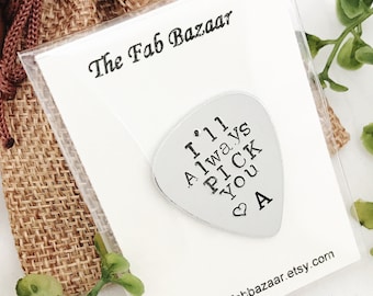 Puede incluir: Una púa de guitarra plateada con el texto "I'll always pick you" y un símbolo de corazón. La púa está en una pequeña bolsa de arpillera con una tarjeta blanca que dice "The Fab Bazaar" y una dirección de sitio web.