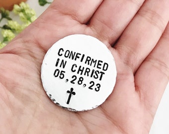 Puede incluir: Un amuleto redondo de plata con el texto "CONFIRMED IN CHRIST 05.28.23" y una pequeña cruz.