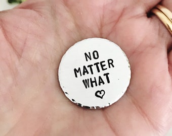 Puede incluir: Un pequeño token redondo de plata con las palabras "NO MATTER WHAT" y un pequeño símbolo de corazón grabado en él.