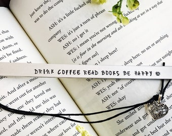 Puede incluir: Un marcapáginas de plata con un cordón negro y un dije de plata. El marcapáginas está grabado con el texto "Drink Coffee Read Books Be Happy".