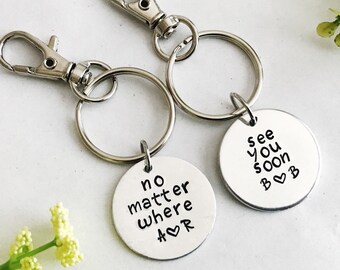 Puede incluir: Dos llaveros plateados con etiquetas redondas de metal. Una etiqueta dice "no matter where" con un símbolo de corazón. La otra etiqueta dice "see you soon" con un símbolo de corazón y la letra "B".
