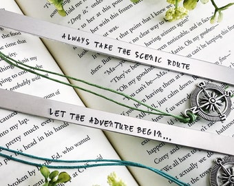 Puede incluir: Dos marcadores de metal plateado con las frases "ALWAYS TAKE THE SCENIC ROUTE" y "LET THE ADVENTURE BEGIN..." se muestran en un libro abierto. Cada marcador tiene un cordón de color y un dije de brújula.