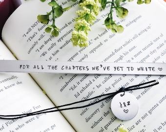 Puede incluir: Un marcapáginas de plata con un cordón negro y un pequeño amuleto de plata con las iniciales "J & Z" y un corazón. El marcapáginas tiene el texto "FOR ALL THE CHAPTERS WE'VE YET TO WRITE" grabado.