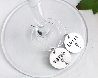 Puede incluir: Dos colgantes plateados para copas de vino con los nombres "Sarah" y "Katie" estampados. Cada colgante tiene un pequeño icono de copa de vino. Los colgantes cuelgan de una copa de vino.