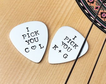 Puede incluir: Dos púas de guitarra blancas con el texto "I PICK YOU" e iniciales. Una púa tiene "C ♡ L" y la otra "K + G". Las púas están sobre una guitarra acústica de madera.