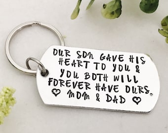 Puede incluir: Un llavero plateado con una etiqueta rectangular. La etiqueta tiene el texto "OUR SON GAVE HIS HEART TO YOU & YOU BOTH WILL FOREVER HAVE OURS. MOM & DAD" en letras negras. El llavero está rodeado de vegetación.