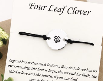 Puede incluir: Una pulsera negra con un colgante redondo con un diseño de trébol de cuatro hojas, presentada en una tarjeta blanca con el texto "Four Leaf Clover". La tarjeta también incluye texto sobre el significado de cada hoja.