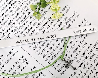 Puede incluir: Un marcapáginas de plata con el texto "WASHED BY THE WATER" y "KATE 09.08.19" grabado en él. El marcapáginas está atado a un cordón verde con un dije de cruz de plata.