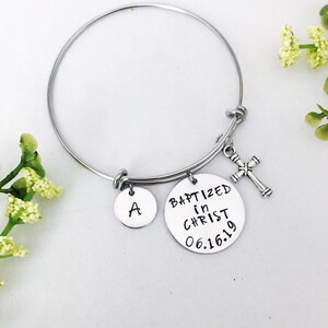 Puede incluir: Un brazalete de plata con un dije que dice "Bautizado en Cristo 06.16.19" y un dije con la letra "A". El brazalete tambi&eacute;n tiene un peque&ntilde;o dije de cruz de plata.