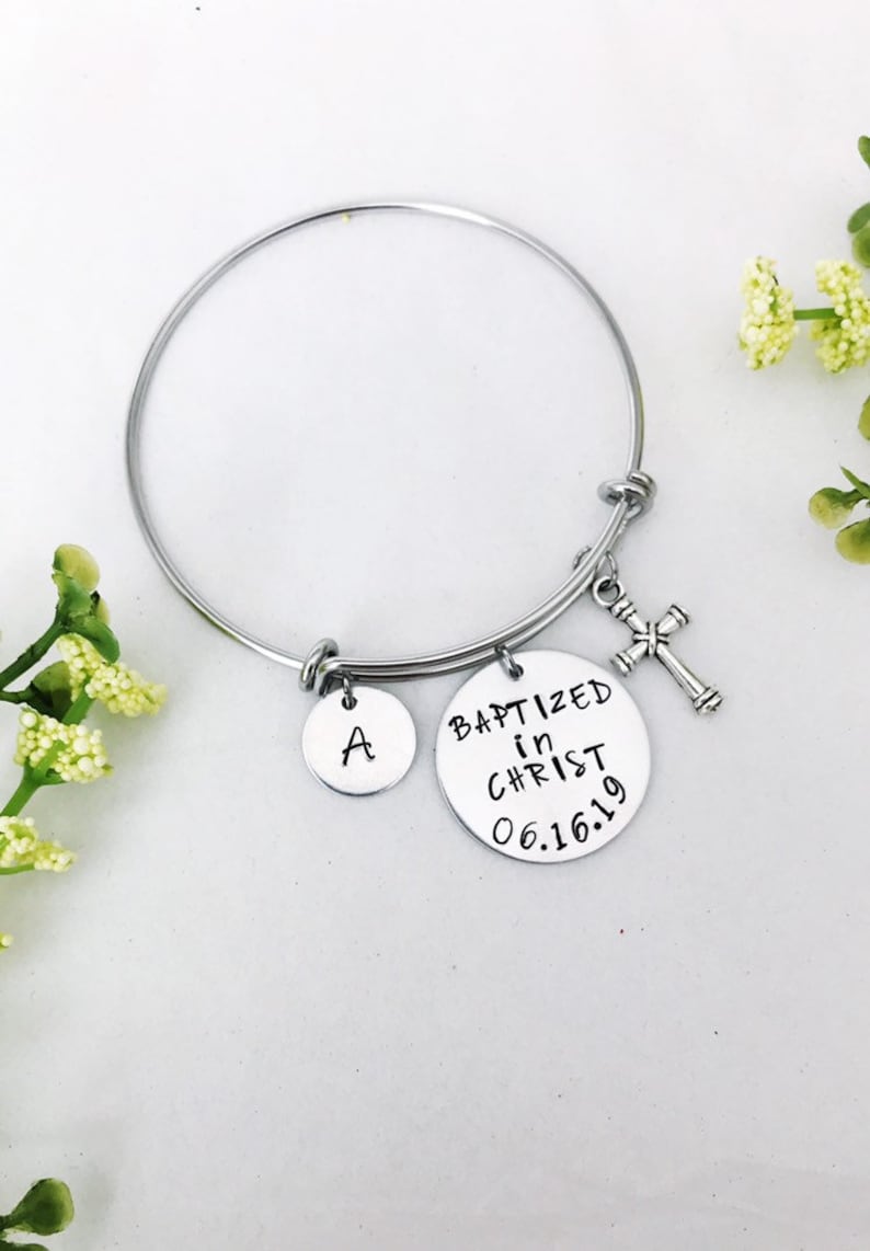 Puede incluir: Un brazalete de plata con un dije que dice "Bautizado en Cristo 06.16.19" y un dije con la letra "A". El brazalete tambi&eacute;n tiene un peque&ntilde;o dije de cruz de plata.